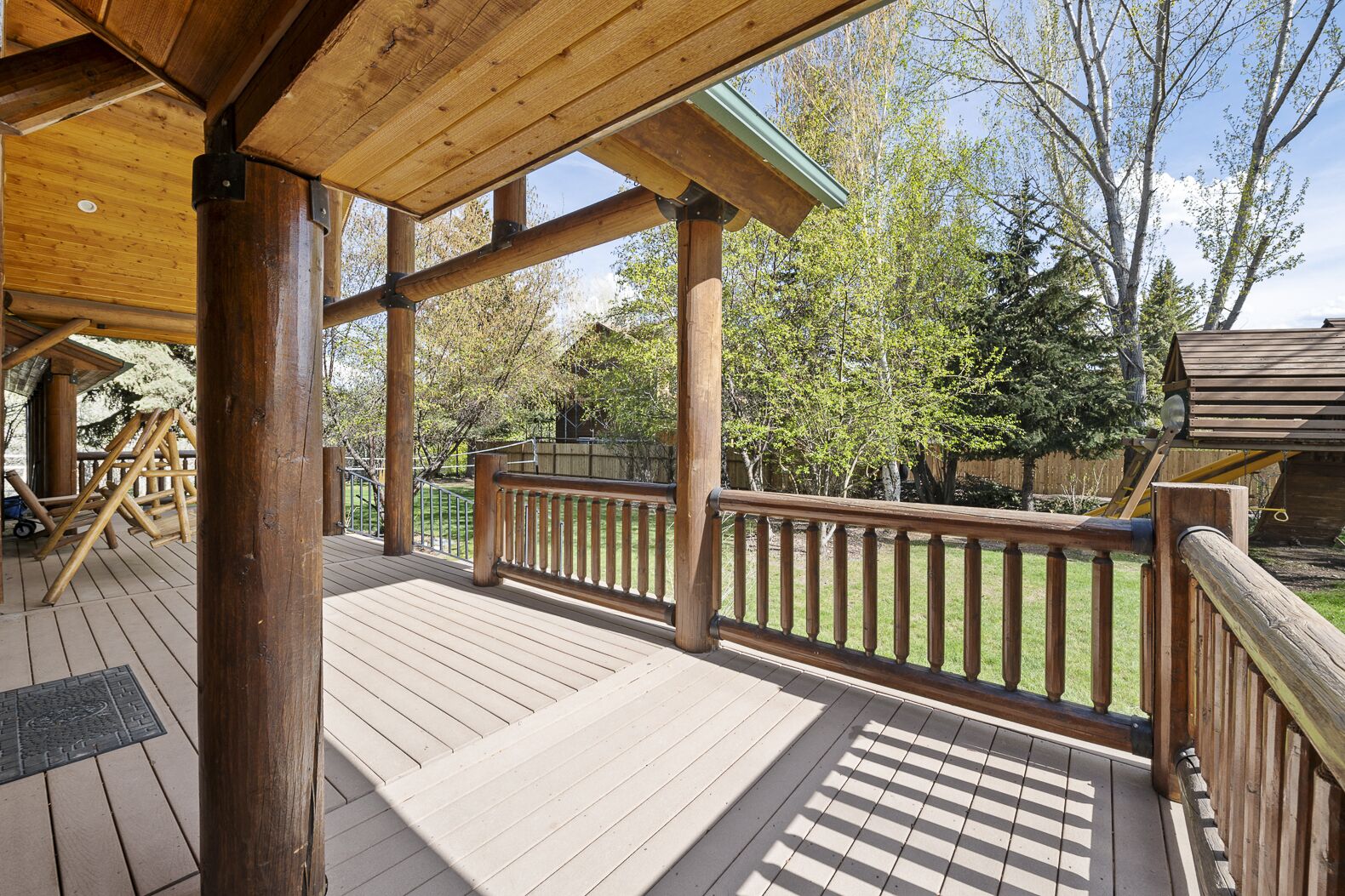 A wooden deck with a railing, a pergola, and a view of trees and a swing set.