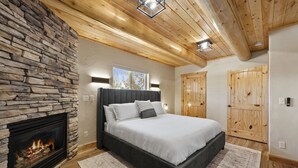 A bedroom with a stone fireplace, a bed with white bedding, wooden ceiling beams, and two wooden doors.