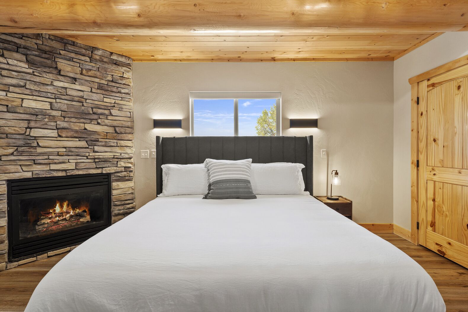 A bedroom with a stone fireplace, a large bed with white bedding, a nightstand with a lamp, and a wooden wardrobe.