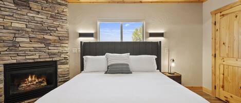 A bedroom with a stone fireplace, a large bed with white bedding, a nightstand with a lamp, and a wooden wardrobe.