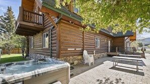 A log cabin with a hot tub, outdoor seating, and a stone fireplace.
