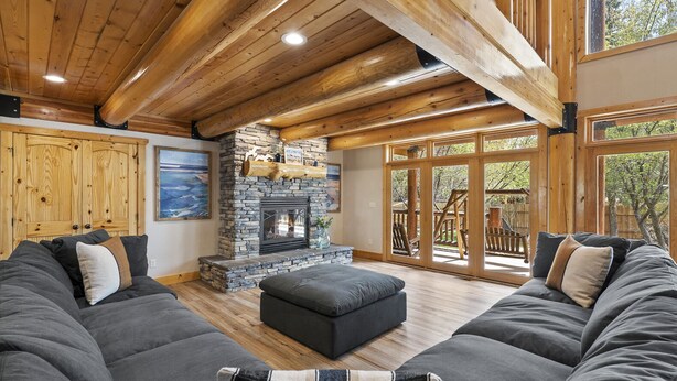 A spacious living room with a stone fireplace, wooden beams, and a large window overlooking a deck.