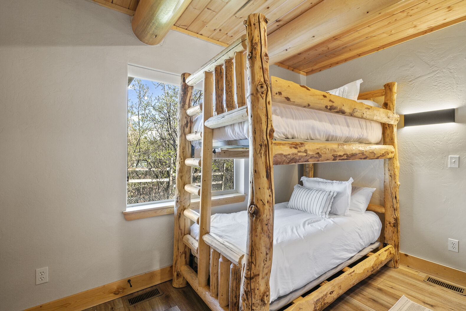 A bunk bed with a wooden frame and white bedding, a window with a view of trees, and a light switch on the wall.