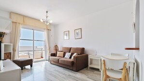 Uma sala de estar moderna com um sofá, uma mesinha e uma cadeira. Há uma janela grande com cortinas, uma luminária no teto e vista para o exterior.