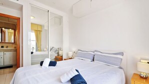 Quarto de hotel com uma cama grande, uma mesinha de cabeceira de madeira, um espelho e um banheiro com pia e espelho.