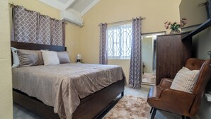A bedroom with a bed, a chair, a wardrobe, a window with curtains, and an air conditioning unit. - Sky View Villas @ Edmund Ridge Estate (Montego Bay)