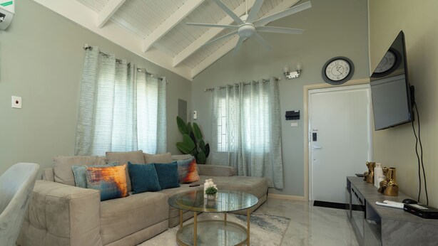 A living room with a sofa, a glass coffee table, a ceiling fan, and a TV mounted on the wall. - Sky View Villas @ Edmund Ridge Estate (Montego Bay)