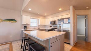 A modern kitchen with white cabinets, a large island with stools, stainless steel appliances, and a window with blinds.