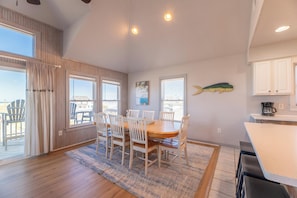A dining area with a wooden table and chairs, a view through large windows, and a kitchen area with white cabinets. - Tapped Out - KDH MP 8, Ocean View, 180 Yds to Beach, Pool, Hot Tub, Pets OK, Flex Stays (Kill Devil Hills)