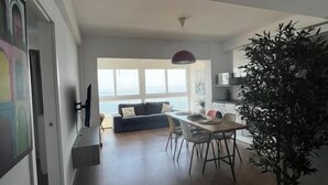 A modern living room with a sofa, dining table, and chairs. There is a large window with a view of the sea.