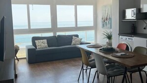 A modern living room with a sofa, dining table, and chairs. A large window offers a view of the sea.