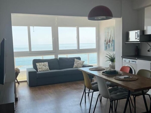 Ein modernes Wohnzimmer mit einer Couch, einem Esstisch und Stühlen. Ein großes Fenster bietet Blick auf das Meer.