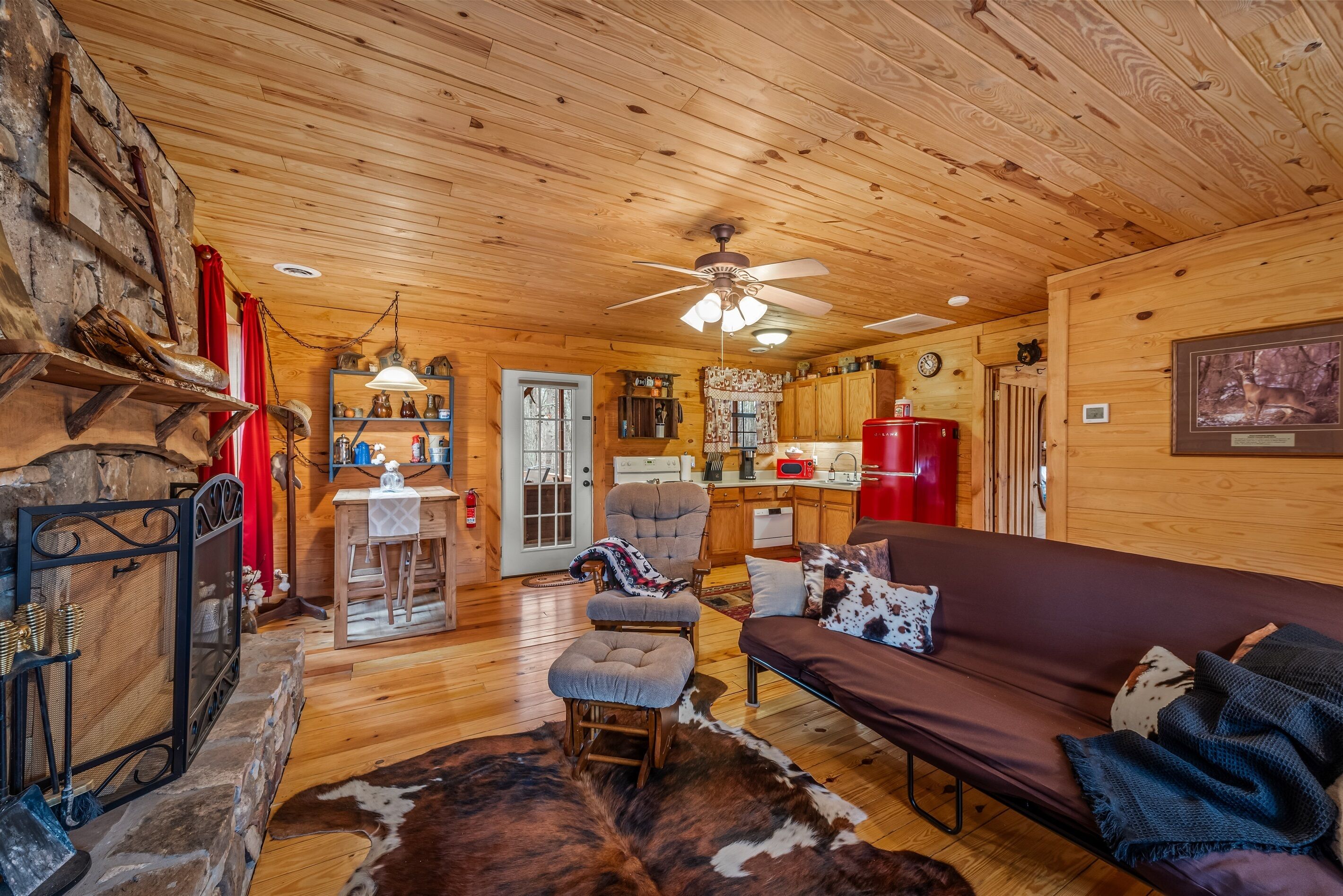 A cozy cabin interior with a stone fireplace, wooden ceiling, a dining area, and a living room with a couch and ottoman.