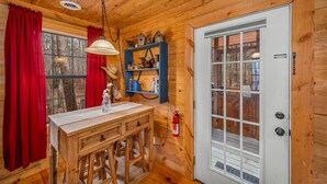 A wooden cabin interior with a dining table, red curtains, a shelf with decorative items, and a white door. - Deerwood-private, cozy cabin in the woods (Mentone)