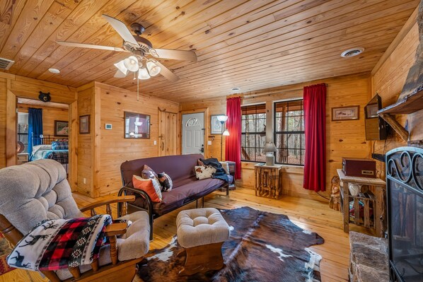 A cozy cabin interior with a wooden ceiling, a sofa, a chair, a small table, and a cowhide rug. - Deerwood-private, cozy cabin in the woods (Mentone)