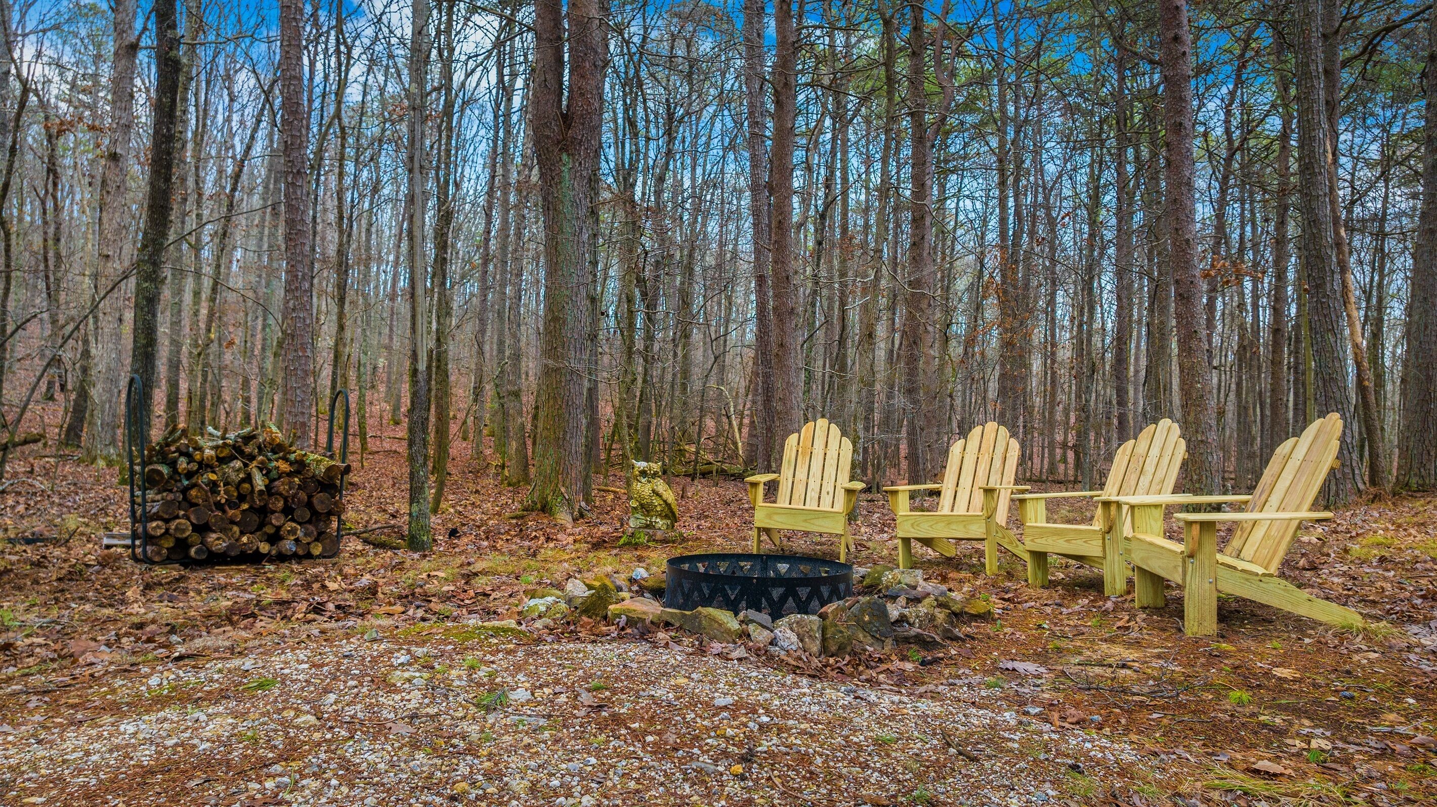 A forest with a pile of logs, a fire pit, and three wooden chairs.