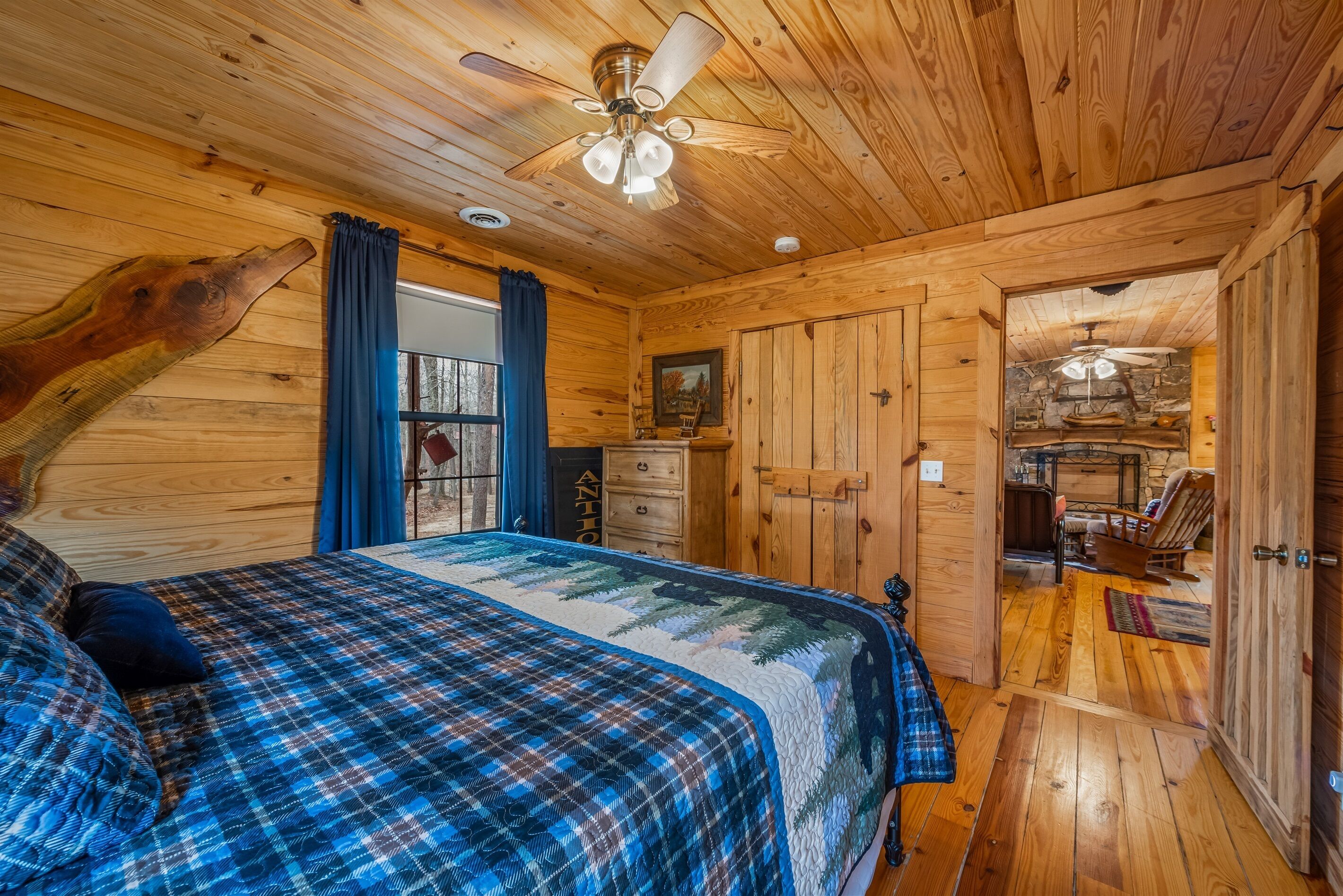 Un dormitorio tipo cabaña de madera con una cama, un ventilador de techo, un aparador y vista hacia la zona de la cocina.