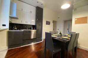 Uma cozinha moderna com eletrodomésticos de aço inoxidável, um espaço de jantar com mesa e cadeiras pretas, e piso de madeira.