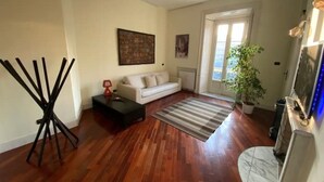 A living room with hardwood flooring, a white sofa, a coffee table, a floor lamp, a painting on the wall, and a potted plant.
