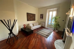 Uma sala de estar com assoalho de madeira, um sofá branco, uma mesa de centro, um abajur, um quadro na parede e um vaso com planta.