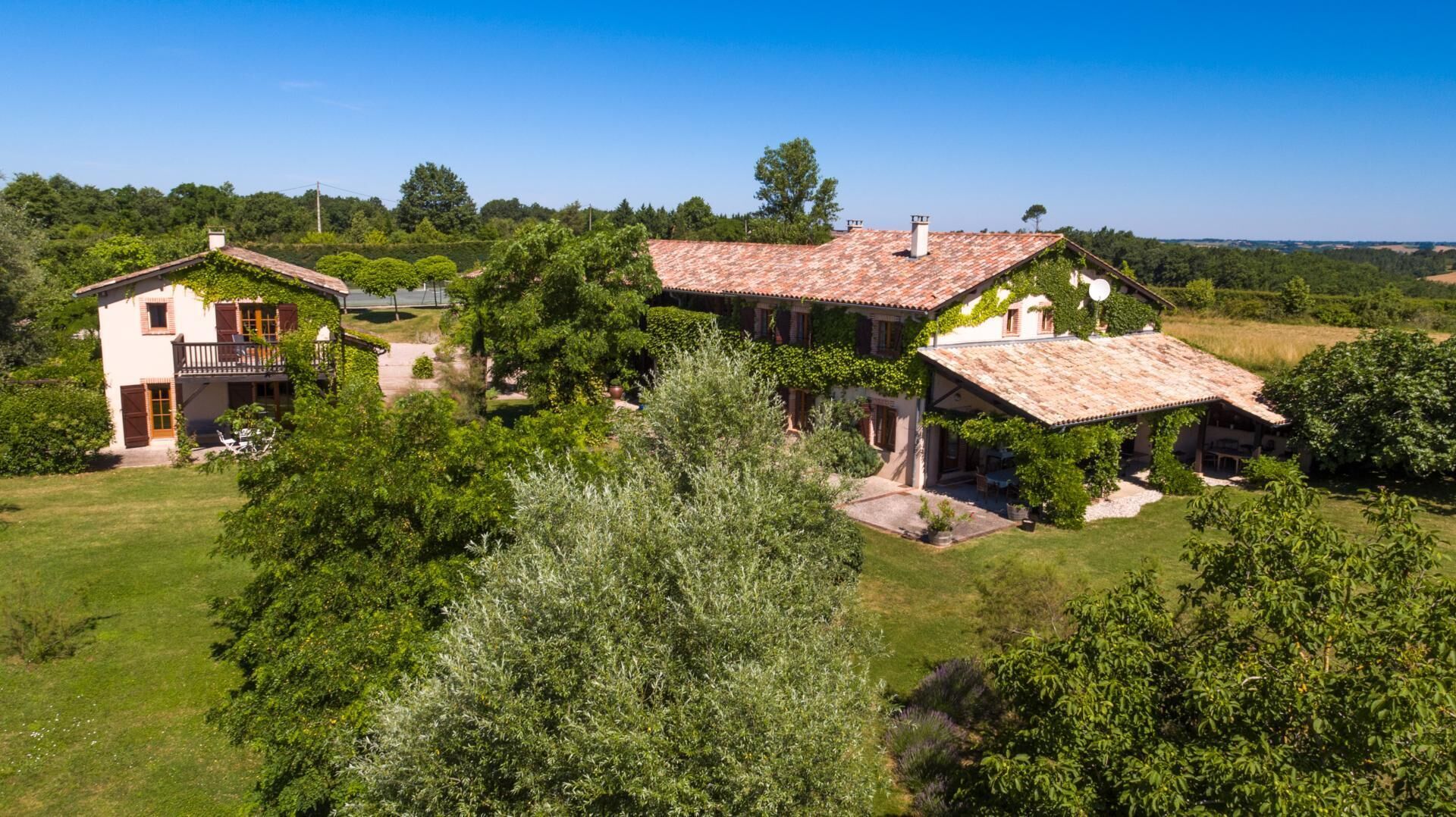 Une maison à deux étages, dotée d’un toit en tuiles, entourée d’une végétation luxuriante et d’arbres.