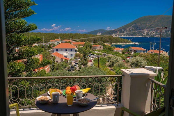 A balcony with a table set for breakfast, overlooking a coastal village with houses and a clear view of the sea.