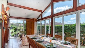 An outdoor dining area with a long table set for a meal, surrounded by large windows and wooden beams.