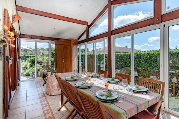 An outdoor dining area with a long table set for a meal, surrounded by large windows and wooden beams.