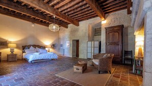 A rustic bedroom with a stone floor, wooden beams, a bed, a wicker chair, and a bookshelf.