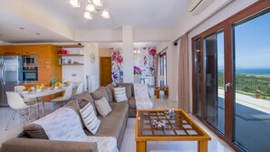 A modern living room with a sofa, coffee table, and dining area. There is a kitchen with appliances and a view of the outdoors. - Iliada - Four Bedroom Villa, Sleeps 8 (Maleme)
