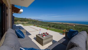 A balcony with a sofa, a table with drinks and fruit, and a view of the sea and hills. - Iliada - Four Bedroom Villa, Sleeps 8 (Maleme)
