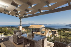 A balcony with wicker furniture, a glass-top table, and a view of the sea and mountains.