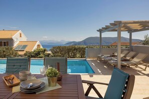 A poolside area with lounge chairs, a table set for dining, and a view of the sea and mountains.