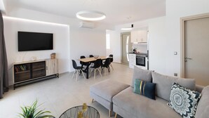 A modern living room with a grey sofa, a dining area with a wooden table and chairs, a kitchen with white cabinets, and a mounted flat-screen TV. - Tina - Three Bedroom Villa, Sleeps 6 (Spartochori)