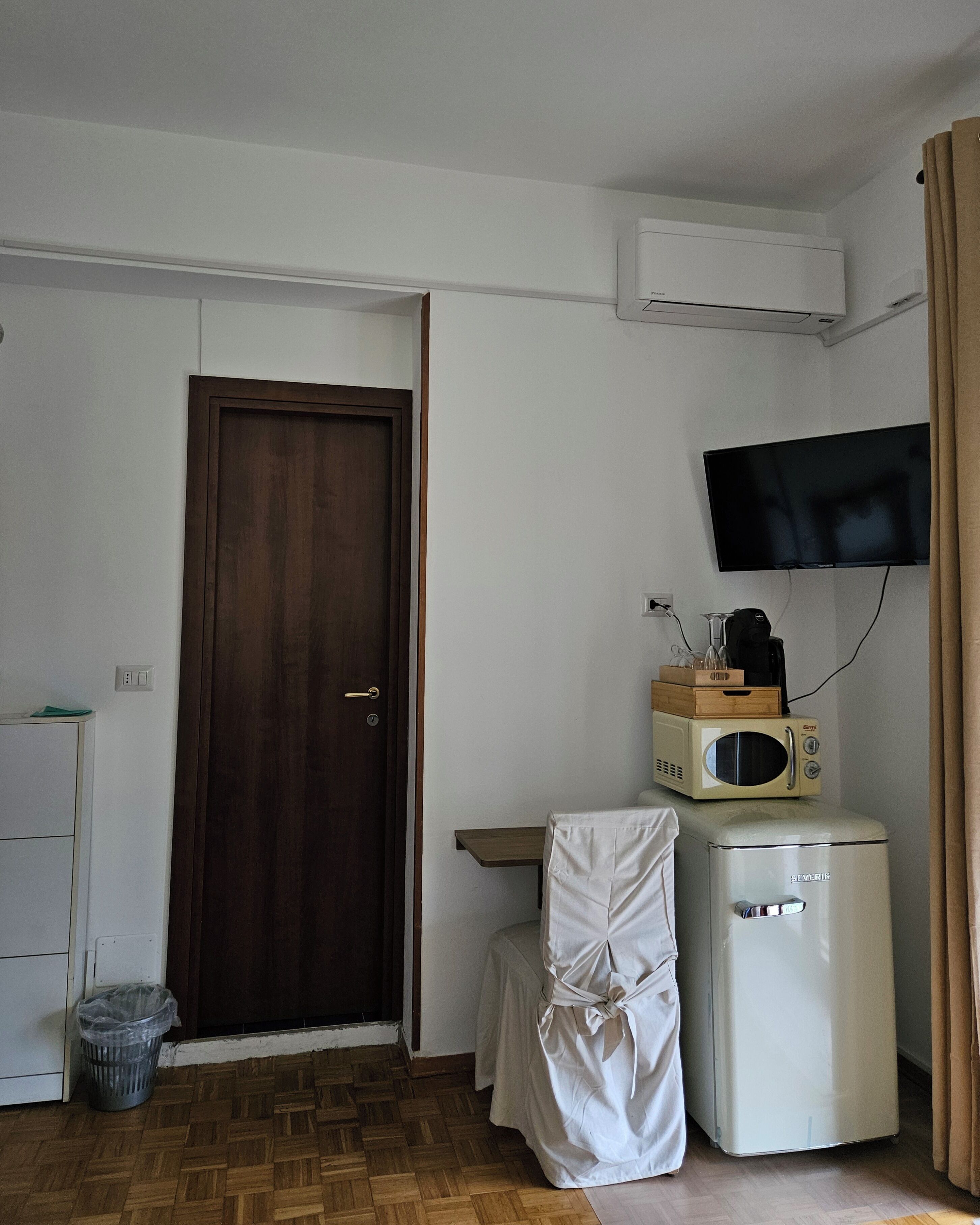 Ein Zimmer mit Holztür, einem weißen Schrank, einem kleinen Tisch mit Sitzauflage, einem weißen Kühlschrank, einer Mikrowelle, einem an der Wand befestigten Fernseher und einem Mülleimer.