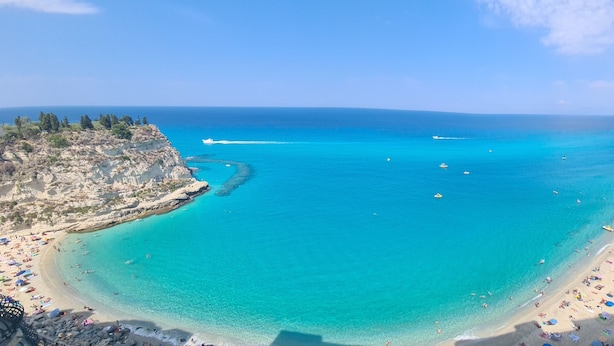 Une plage côtière aux eaux turquoise et limpides, bordée d’une côte rocheuse et dotée d’une plage de sable où l’on trouve des parasols et des gens.