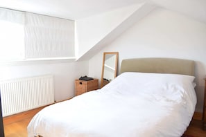 Une chambre avec un plafond en pente, un lit avec une literie blanche, une tête de lit en bois, un petit panier en osier et un miroir sur le mur.