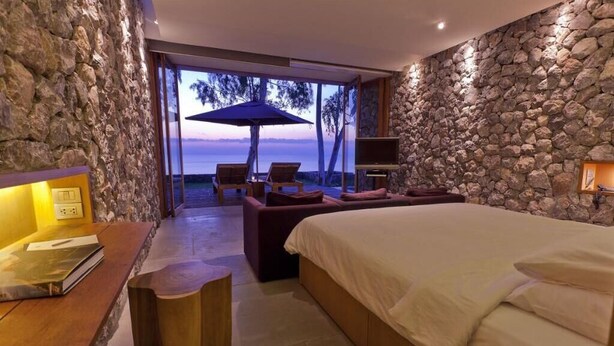 A bedroom with a large bed, a desk with a chair, a sofa, and a TV. There is a stone wall and a view of the ocean.