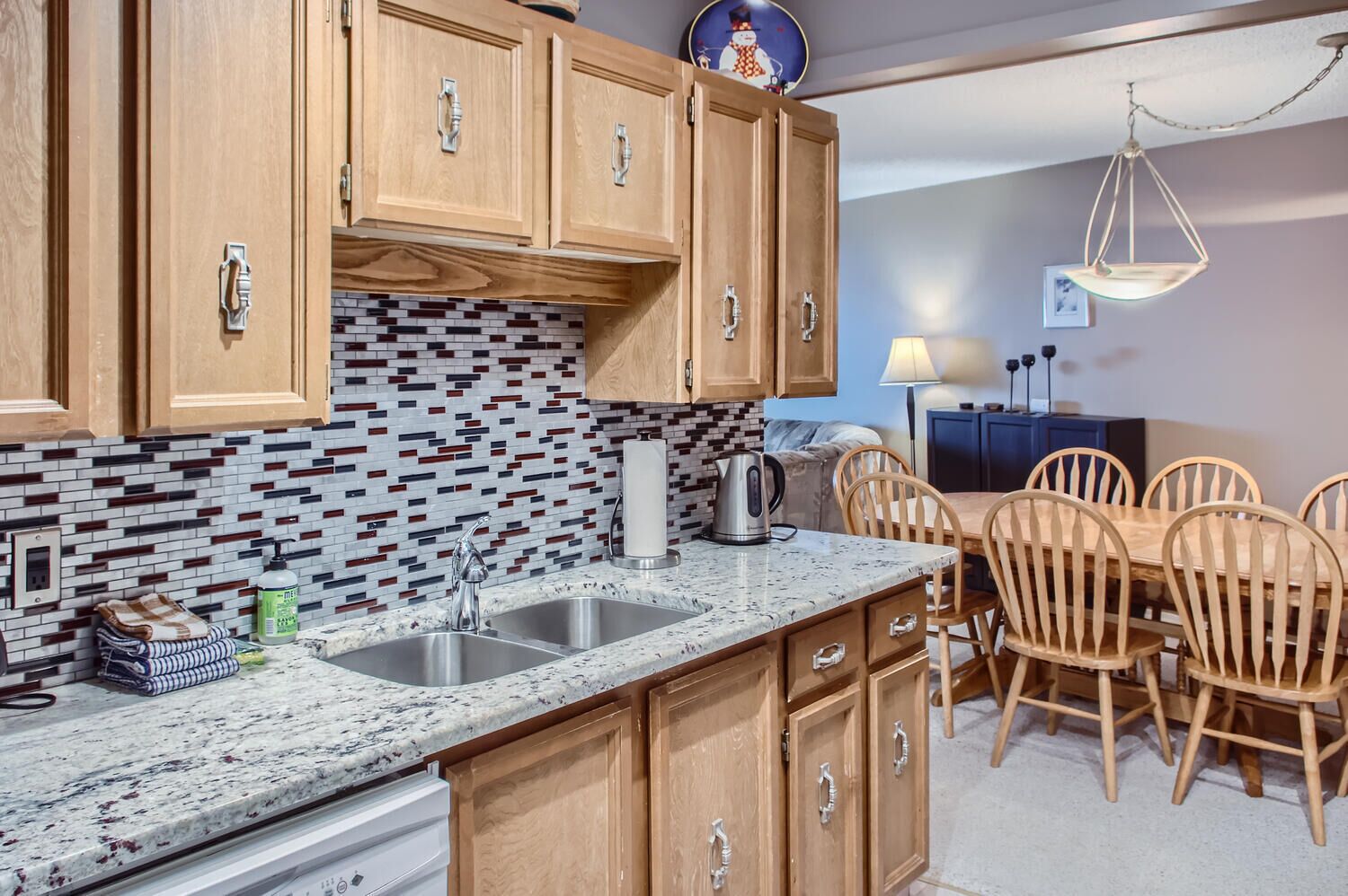 A kitchen with wooden cabinets, a tiled backsplash, a granite countertop, and a dining area with wooden chairs and a table.
