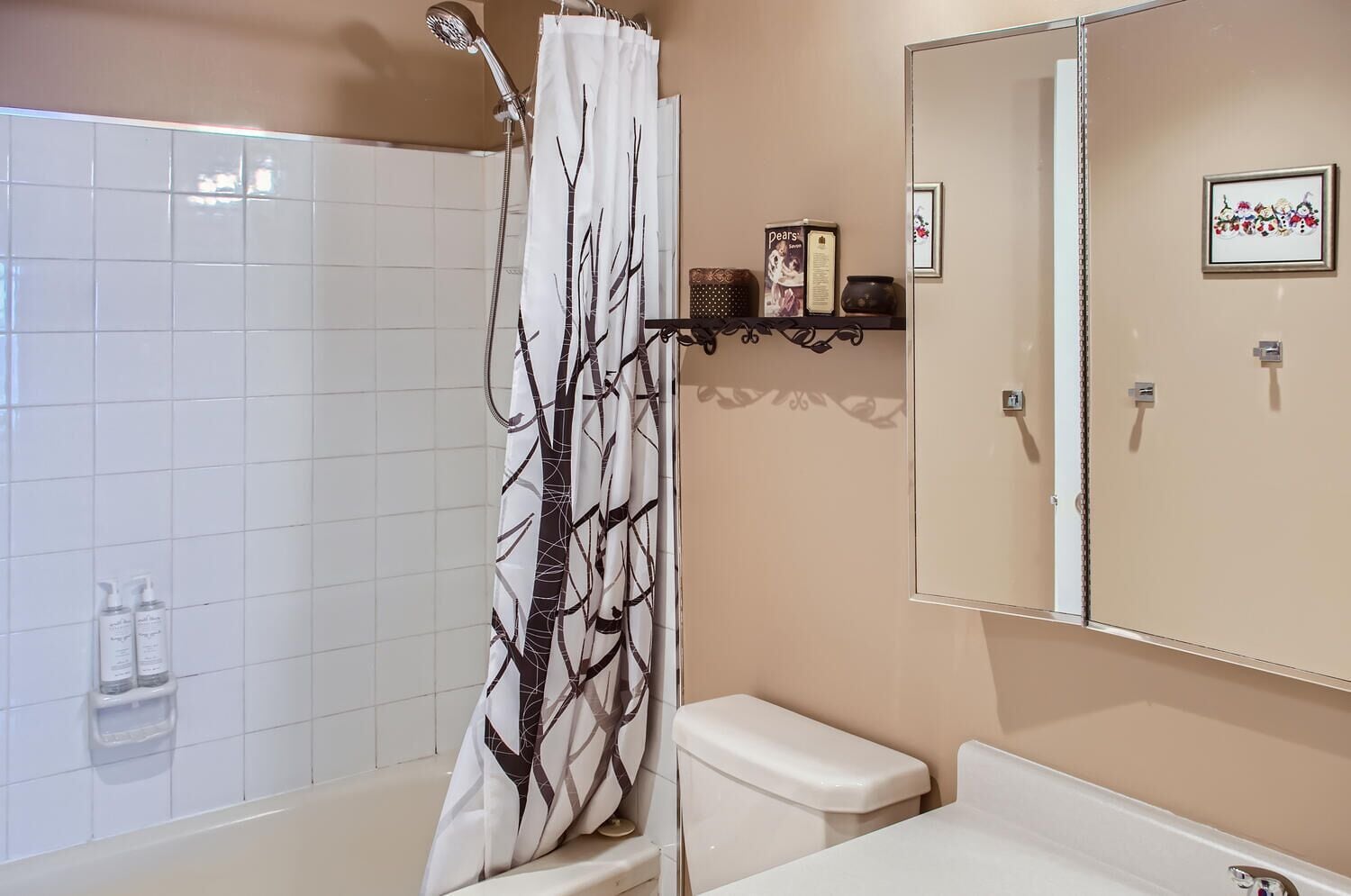 A bathroom with a shower, toilet, and sink. There is a shower curtain with a tree pattern, a mirror above the sink, and a shelf with toiletries.