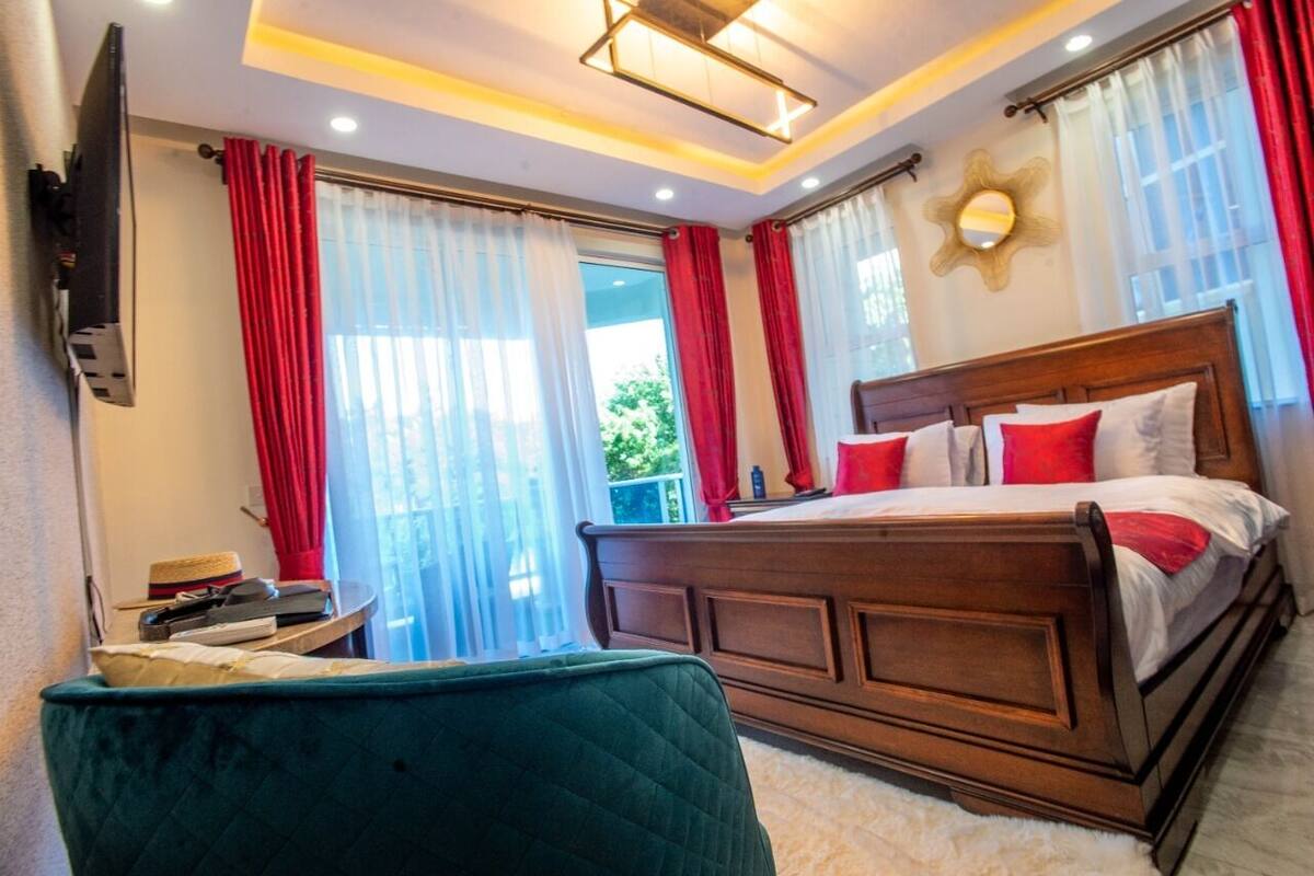 Ein Schlafzimmer mit einem hölzernen Bett, roten und weißen Vorhängen, einem grünen Sessel und einem an der Wand befestigten Fernseher.