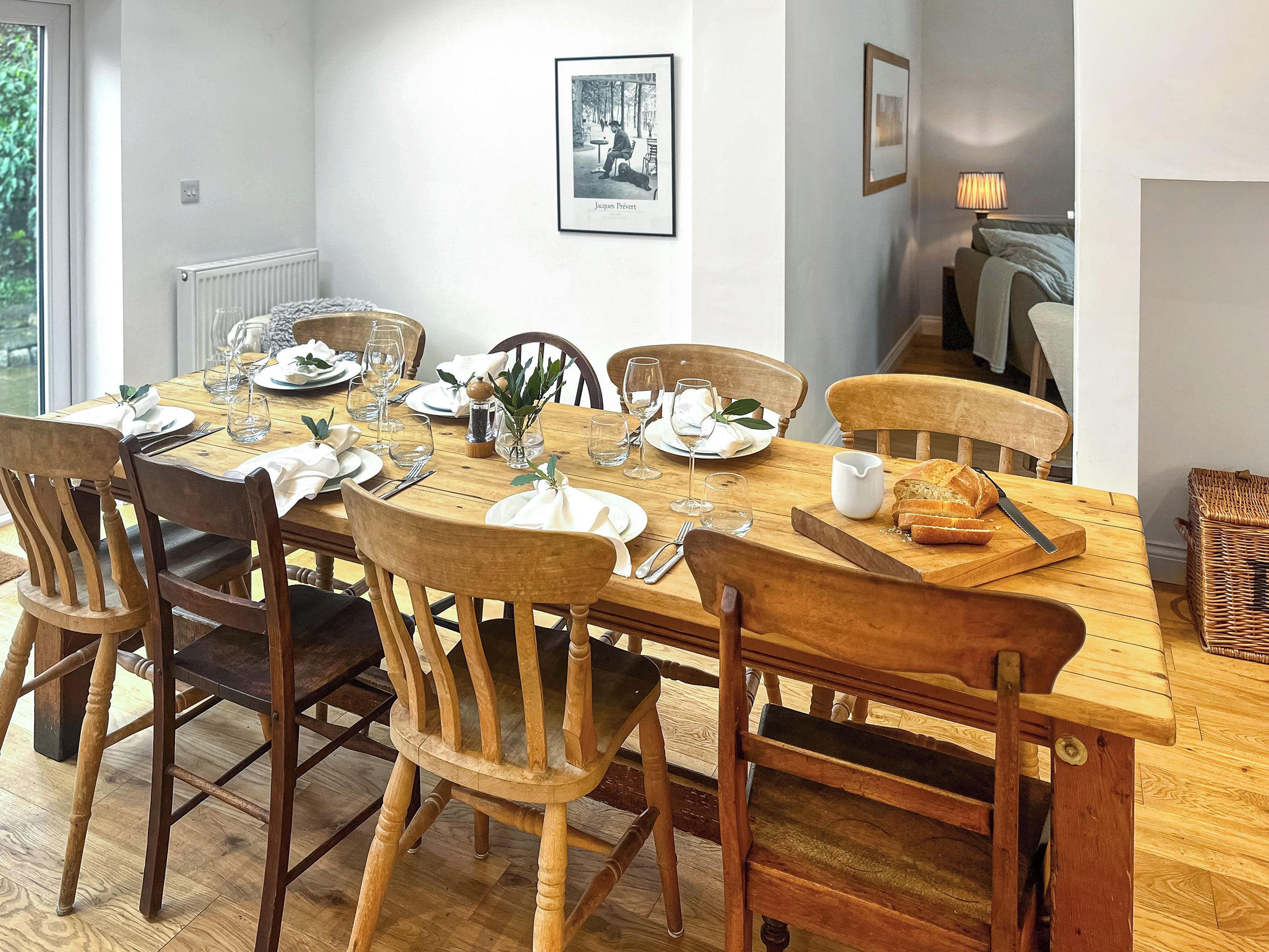 Une table à manger dressée pour six personnes, avec des chaises en bois, un tableau encadré au mur et un panier en osier posé sur le sol.