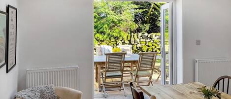 Una habitación con una mesa de comedor de madera, dos sillas, un sofá y vista a un jardín exterior por una puerta abierta.