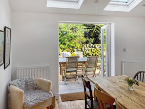 A room with a wooden dining table, two chairs, a sofa, and a view of an outdoor garden through an open door.