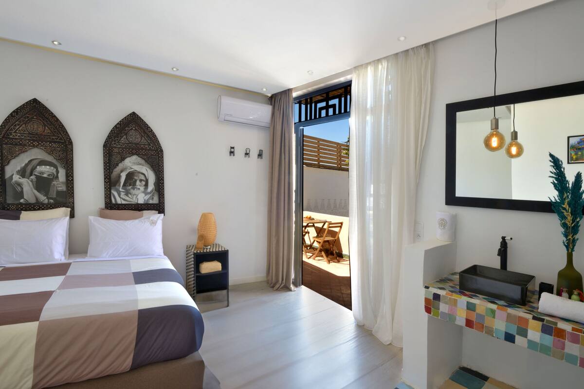 A modern hotel room with a large bed, a small desk, a bathroom with a mirror and sink, and a balcony with a view.