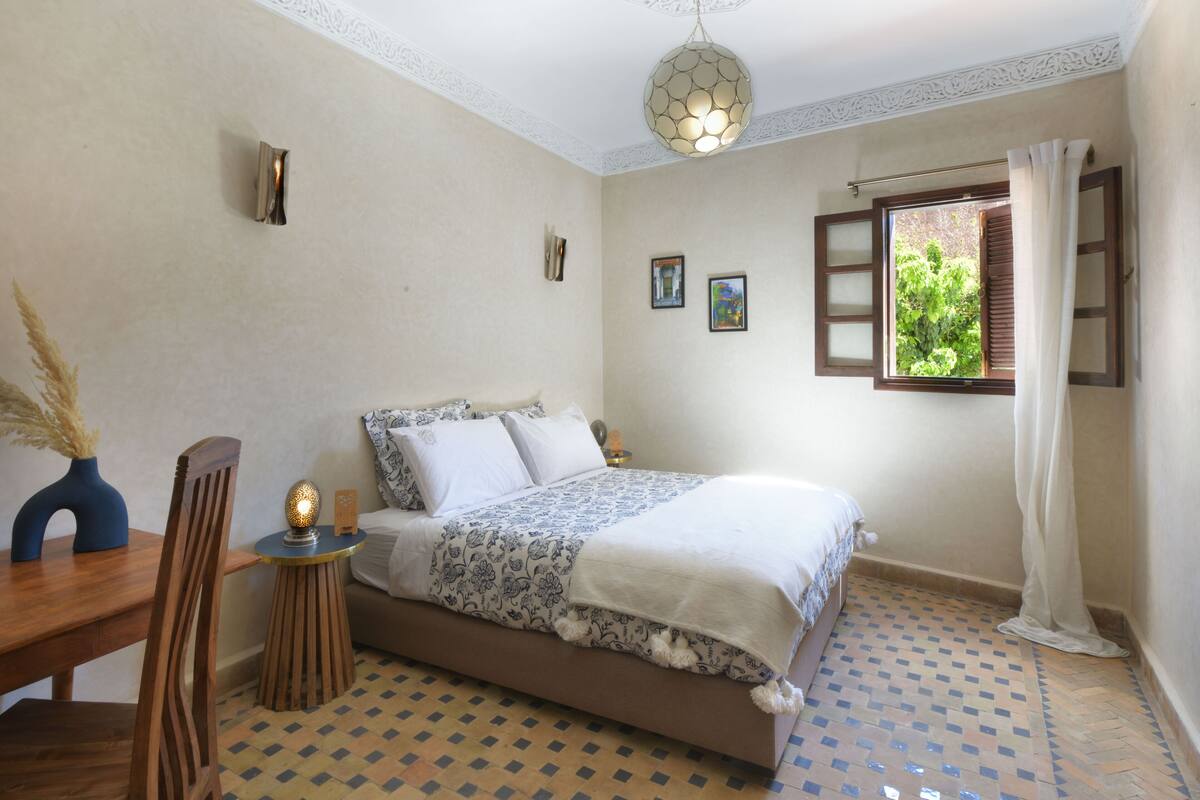 A bedroom with a bed, a small round table, a chair, a lamp, and a window with curtains.