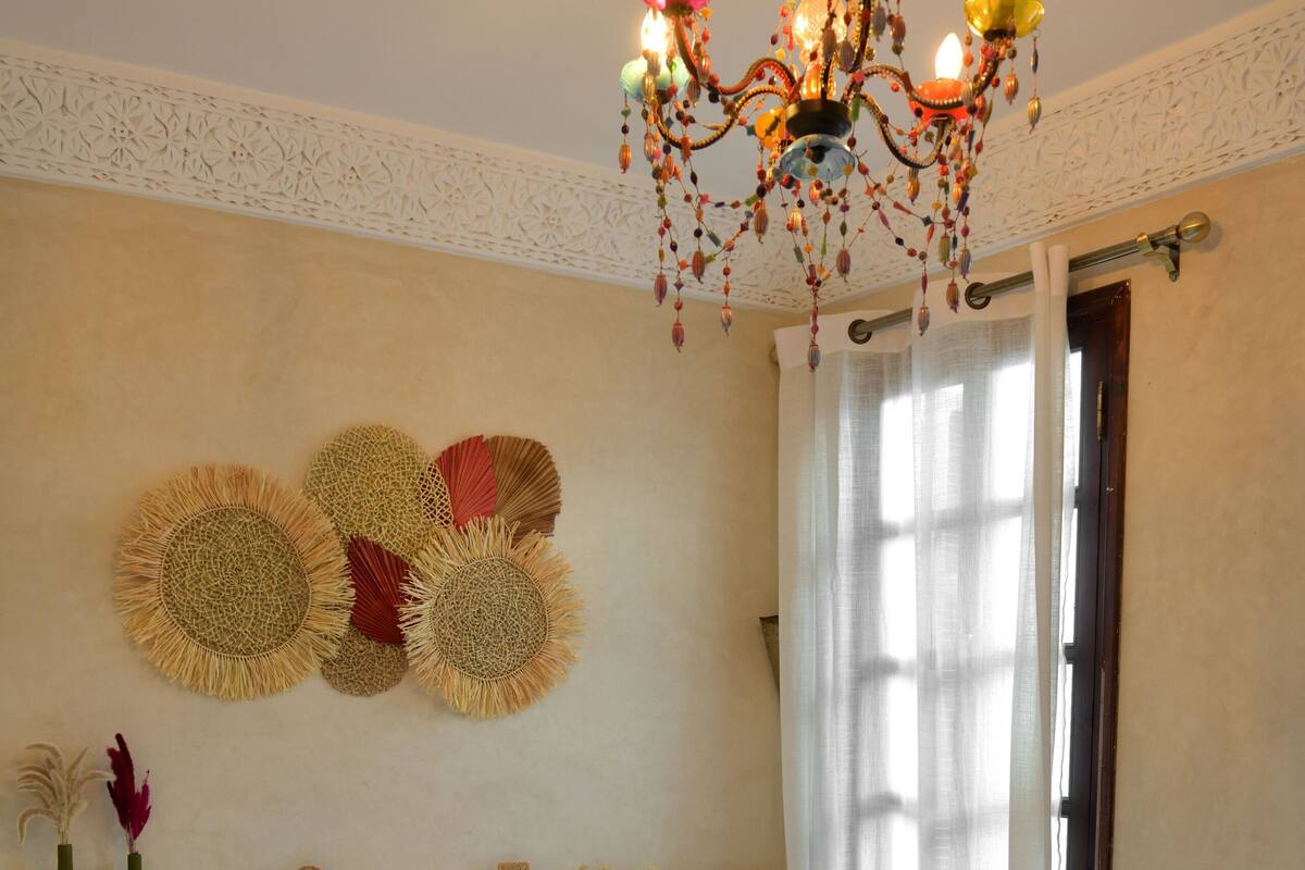 A hotel room with two beds, a chandelier, decorative wall hangings, and a window with curtains.