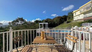 A wooden deck with a table and benches, overlooking a swimming pool and a white gazebo.