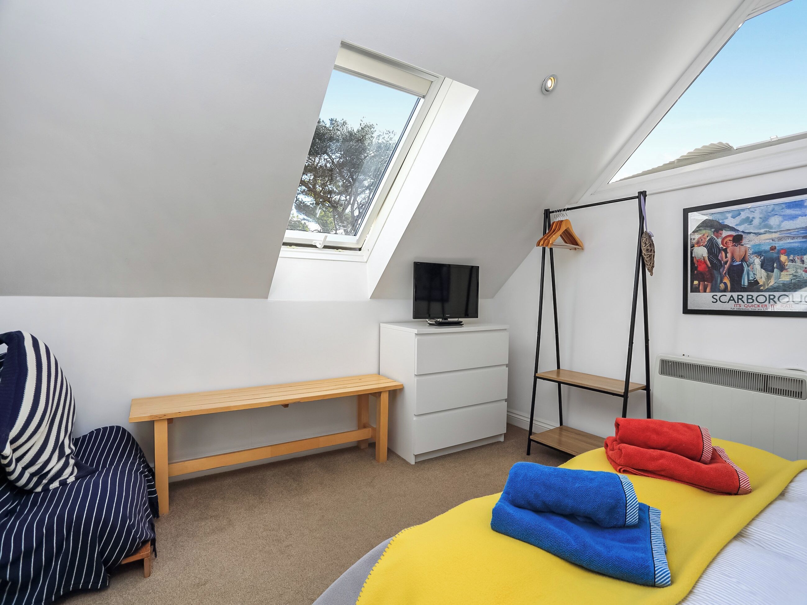 A bedroom with a skylight, a bed with towels, a bench, a TV, a coat rack, and a poster on the wall.