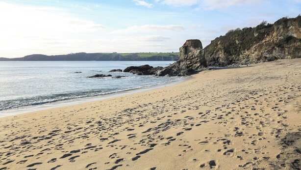 A sandy beach with footprints, a rocky cliff, and calm water.